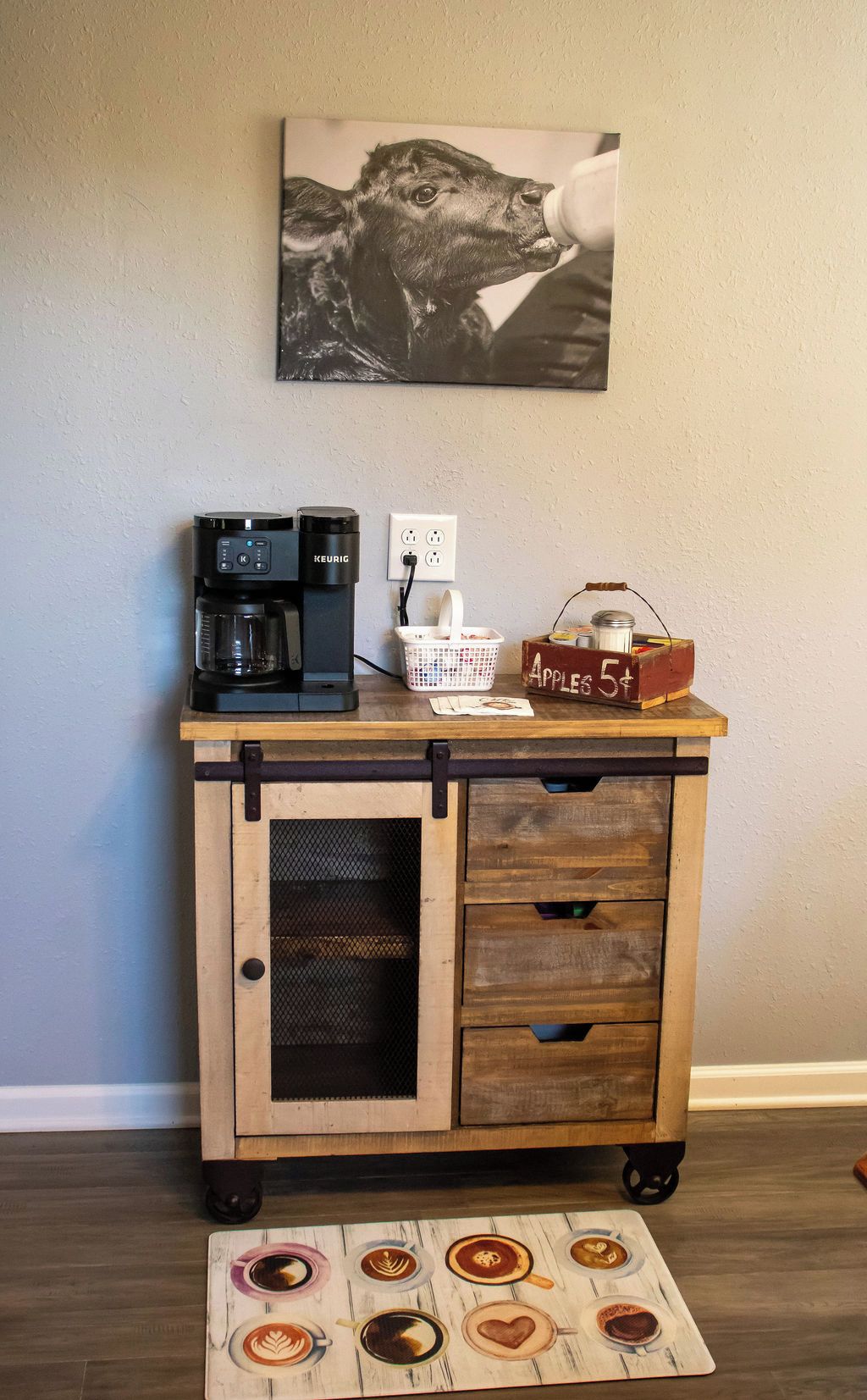 A coffee station with a Keurig machine, a basket, a milk container, and a sign indicating apple prices.