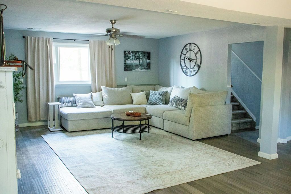 Un salon avec un canapé d’angle, une table basse, une horloge au mur et un escalier sur le côté droit.