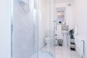 Un baño moderno con ducha envuelta en vidrio, inodoro blanco, lavamanos con resguardo de mosaicos y una planta pequeña sobre la encimera.