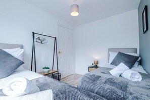 Un dormitorio moderno con dos camas, una mesita de noche con lámpara, una planta pequeña y una puerta.