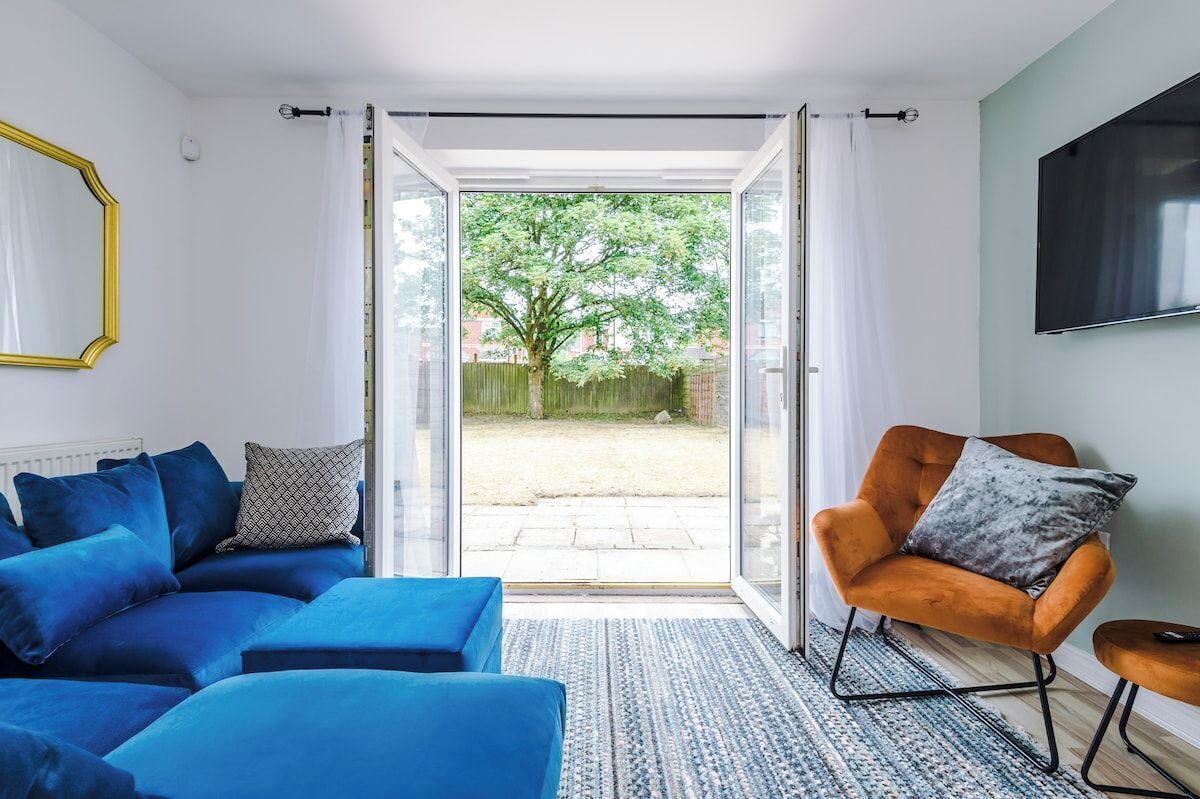 A modern living room with a blue sofa, a patterned throw pillow, a large flat-screen TV, and a sliding glass door leading to a garden.