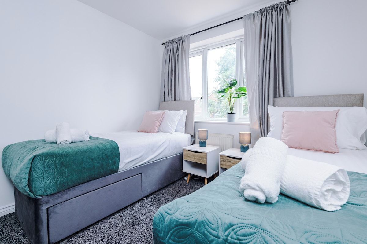 A twin room with two beds, a window with curtains, bedside tables with lamps, and a potted plant.