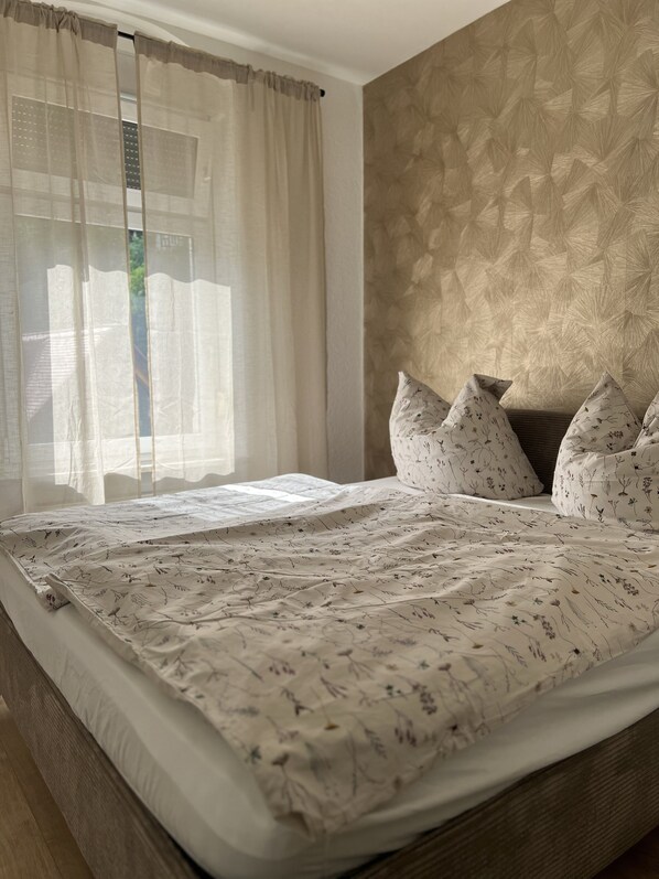 Una cama bien tendida con ropa de cama floral, una ventana con cortinas translúcidas y una pared con textura.