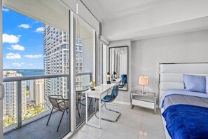 A modern hotel room with a balcony, a desk with a chair, a bed, and a nightstand with a lamp. - High-Floor 1BR | Bay & City Views | Brickell (Miami)
