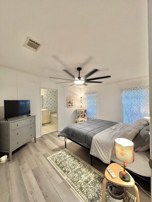 Un dormitorio con cama, televisor sobre una cómoda, ventilador de techo y mesita de noche con lámpara.