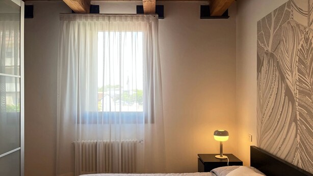 Un dormitorio con una cama, una mesita de noche, una lámpara, una ventana con cortinas translúcidas y una pared con diseño de hojas.