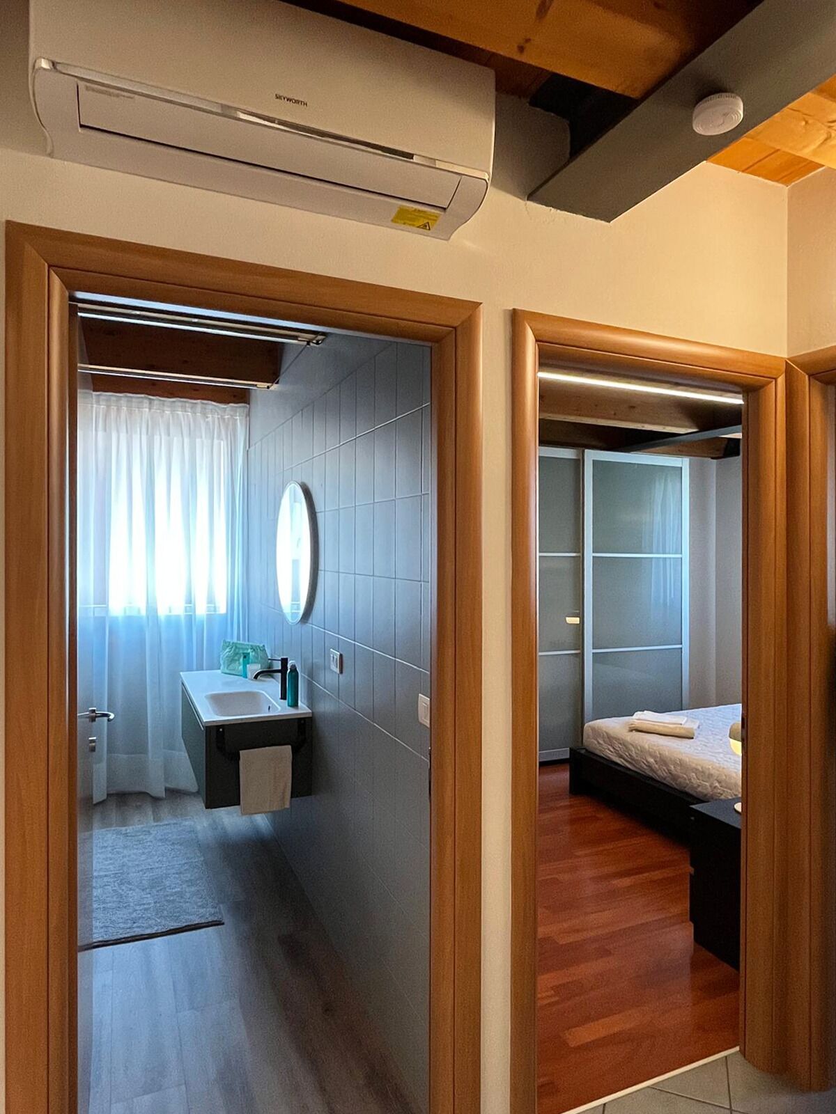 A hotel room with a wooden floor, a bed, a bedside table, a mirror, and a window with curtains.