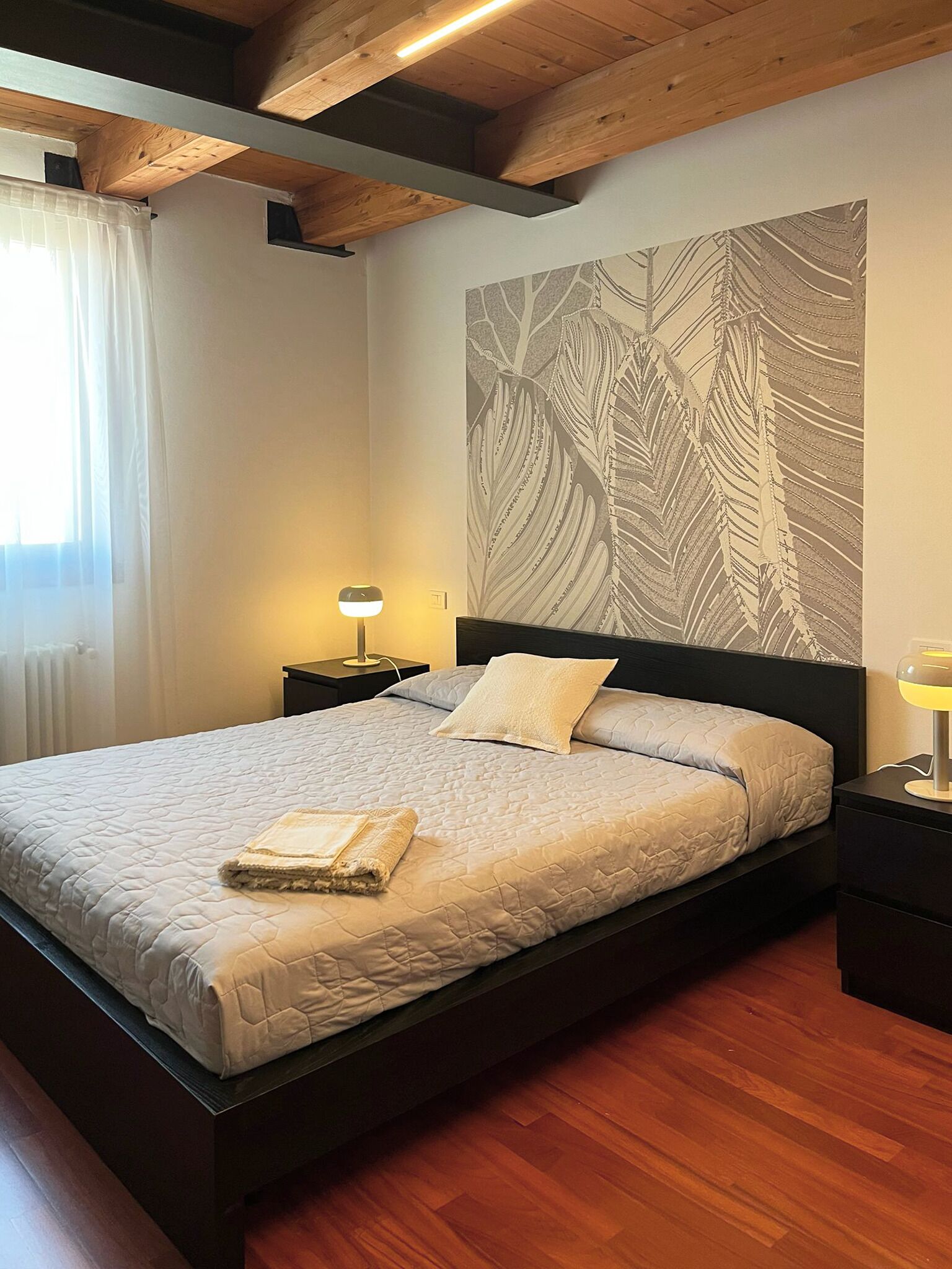 Un lit bien fait, avec une courtepointe, une lampe de chevet et un mur orné d’un motif de feuilles.