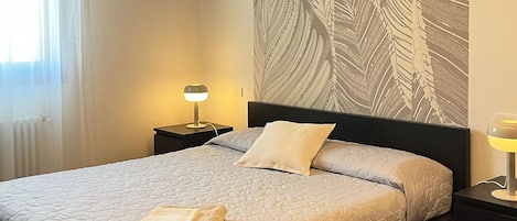 Una cama bien hecha con colcha, una lámpara de noche y una pared con estampado de hojas.
