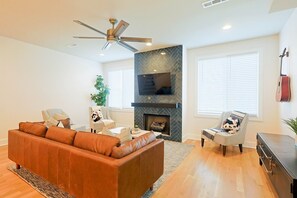 A living room with a fireplace, a sofa, two armchairs, a ceiling fan, and a television mounted on the wall. - Luxury East Nashville Dual Townhomes | Walk to Five Points | 8 Bedrooms |2 Arcade Lounges (Nashville)