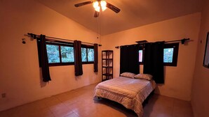 A bedroom with a bed, a ceiling fan, a window with curtains, and a small shelf. - House with a spacious yard surrounded by jungle and trees, ideal for families (Xpujil)