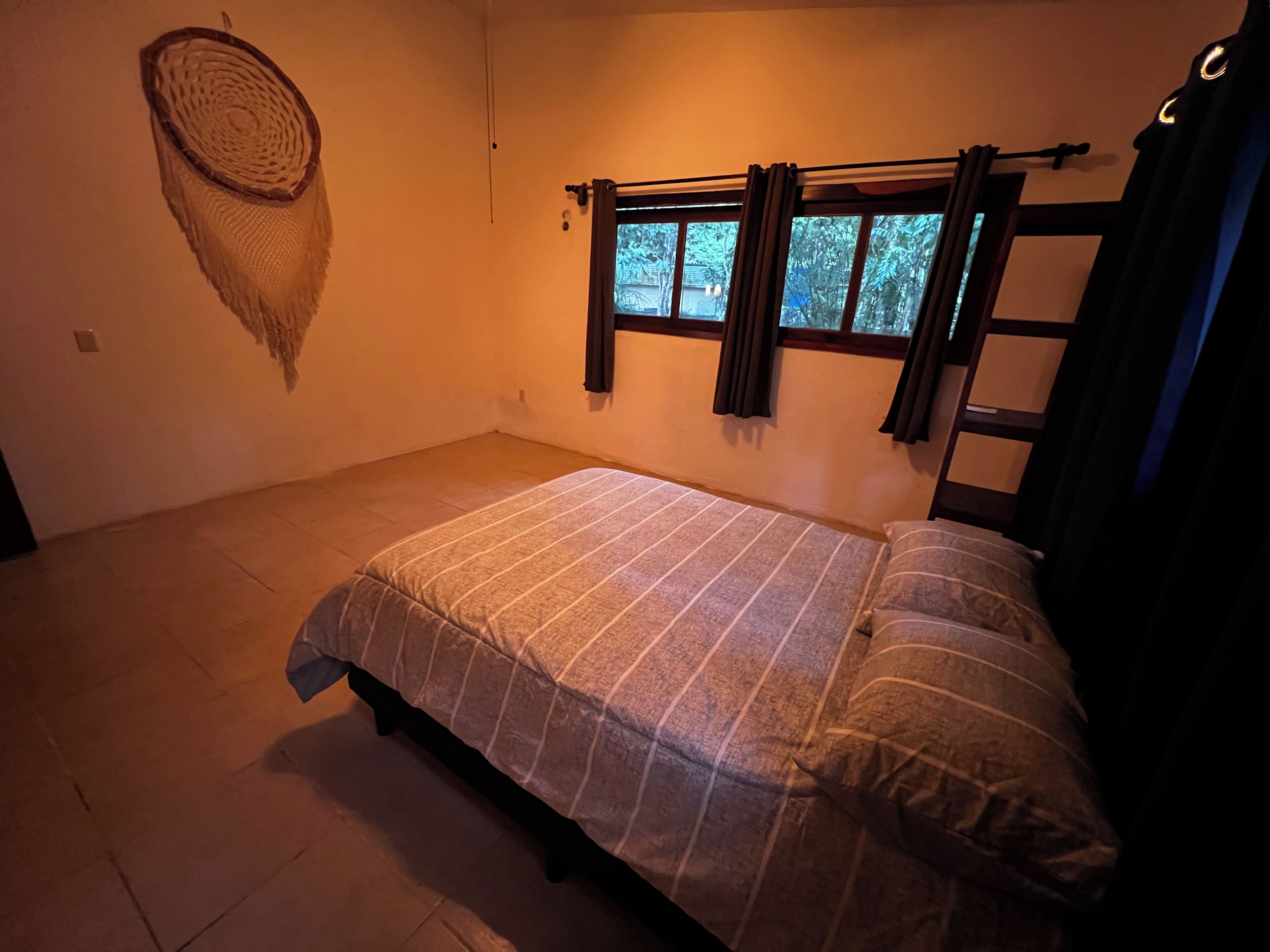A bedroom with a bed, a window with curtains, a woven wall hanging, and a tiled floor.