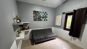 A room with a grey sofa, a tiled floor, a ceiling fan, a window with curtains, and a wall with a decorative mirror and a framed picture. - House with a spacious yard surrounded by jungle and trees, ideal for families (Xpujil)