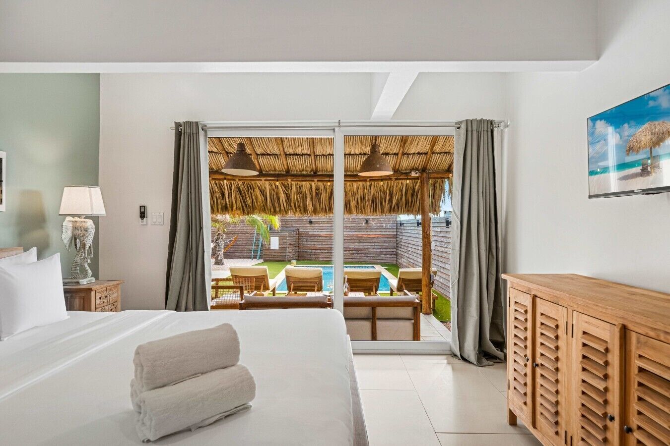 Une chambre d’hôtel avec un lit, une armoire en bois, un bureau avec une lampe et une vue sur l’espace piscine extérieure, visible à travers une porte vitrée.