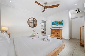 Une chambre d’hôtel avec un lit, un ventilateur de plafond, une commode en bois, un tableau et une petite table avec des serviettes.