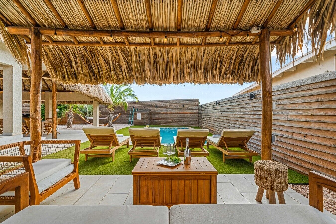 Un espace extérieur aménagé avec une zone de repos couverte, du mobilier en bois, une piscine et une clôture en bois.