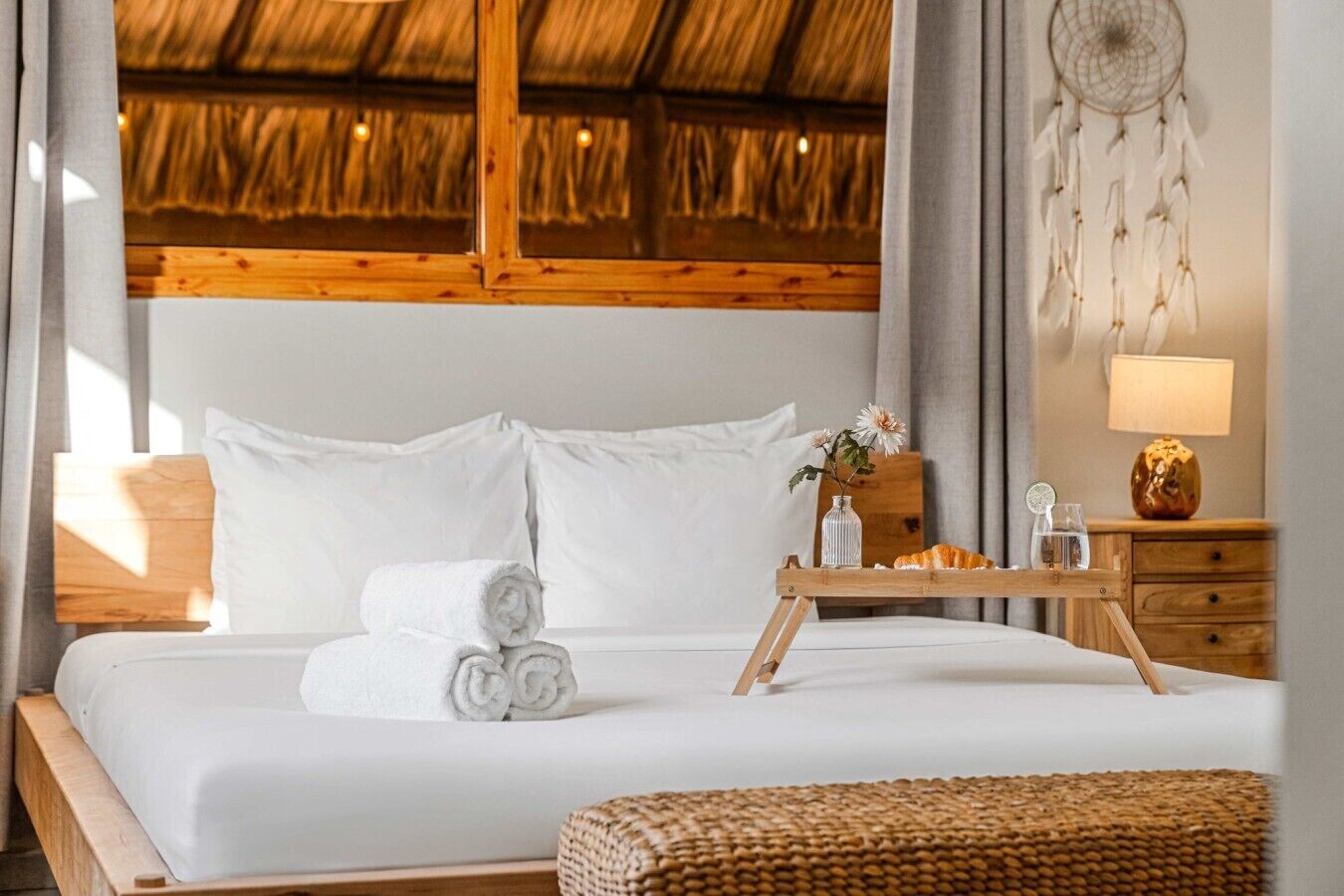 Een net opgemaakt bed met witte lakens en opgerolde handdoeken, een nachtkastje met een lamp, een dromenvanger, en een dienblad met een croissant en een glas water.
