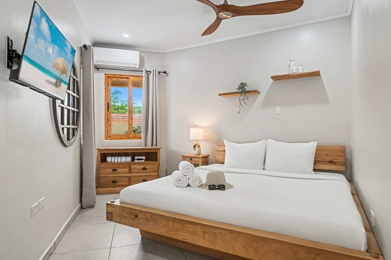 Un dormitorio con cama, ventilador de techo, televisor, ventana con cortinas, cómoda de madera y estante montado en la pared.