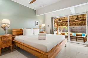 A hotel room with a large bed, wooden headboard, two bedside tables with lamps, a view of an outdoor area with a thatched roof and seating. - 6BR Aruba Villa w/ Pool | Near Palm & Eagle Beach (Noord)