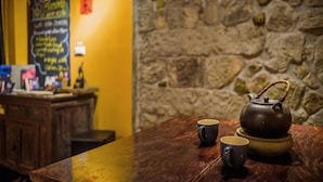 Uma mesa de madeira com um bule e duas xícaras, uma parede de pedra e um balcão com um cardápio em lousa ao fundo.