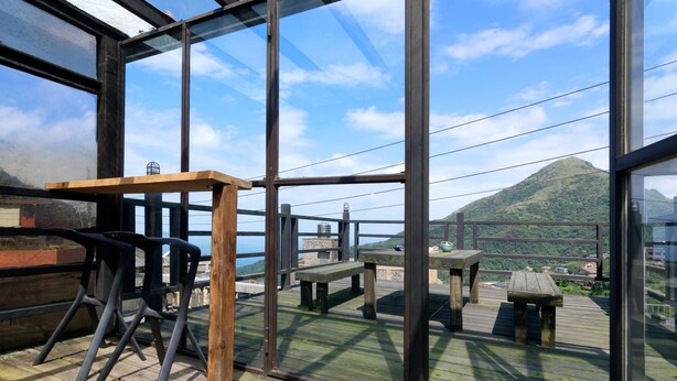Um terraço com mobiliário de madeira e paredes de vidro, oferecendo vista para uma paisagem montanhosa.