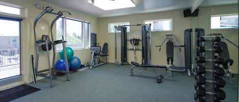 Un gimnasio con equipo de ejercicio, ventilador de techo y amplias ventanas.