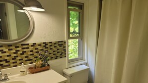 Une salle de bain avec un lavabo blanc, un miroir, une fenêtre donnant sur un espace verdoyant, des toilettes et un rideau de douche.