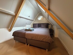 Un dormitorio con techo inclinado, una cama con cabecera, una mesita de noche, una lámpara y un ventanal circular.