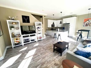 Una sala de estar moderna con televisor, centro de entretenimiento y sofá. Hay una zona de cocina con microondas y refrigerador.