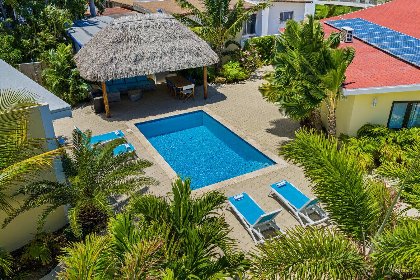 Une piscine entourée de chaises longues, d’une structure à toit de chaume et d’une végétation tropicale luxuriante.