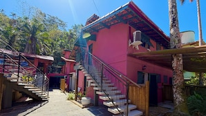 A pink building with a staircase leading to a second floor, surrounded by greenery and a clear sky.