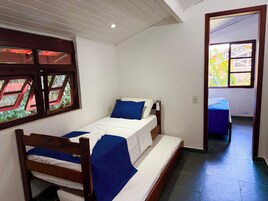 Cama de solteiro com estrutura de madeira, roupa de cama branca e detalhes em azul. Uma janela mostra vista para o verde.