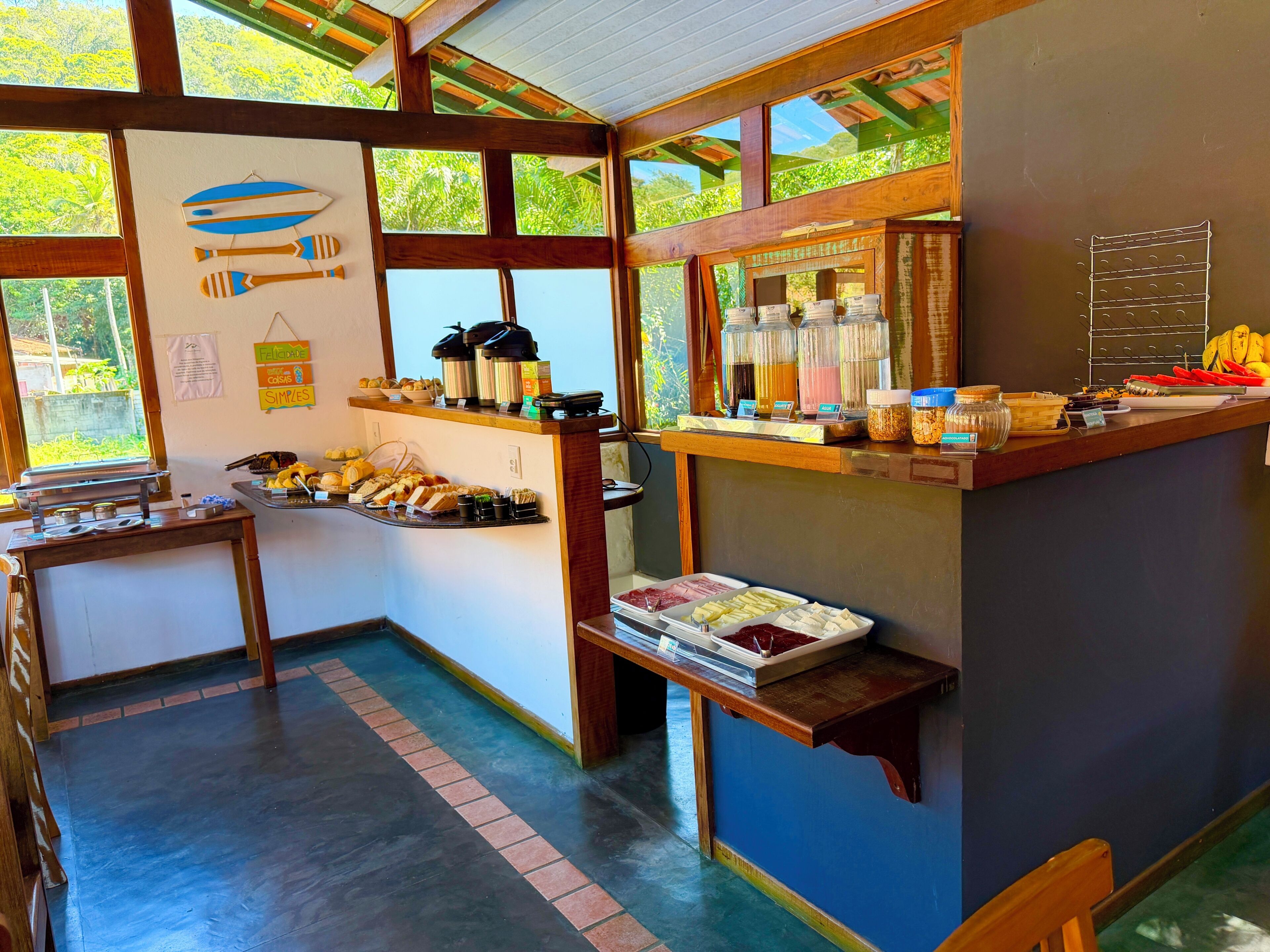 A breakfast bar with various food items and beverages, a wall with hanging decorations, and a view of greenery through large windows.