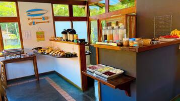 A breakfast bar with various food items and beverages, a wall with hanging decorations, and a view of greenery through large windows.