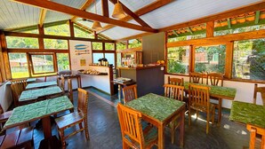A restaurant with wooden tables and chairs, a counter with food items, and large windows offering a view of greenery.