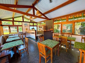 A restaurant with wooden tables and chairs, a counter with food items, and large windows offering a view of greenery.