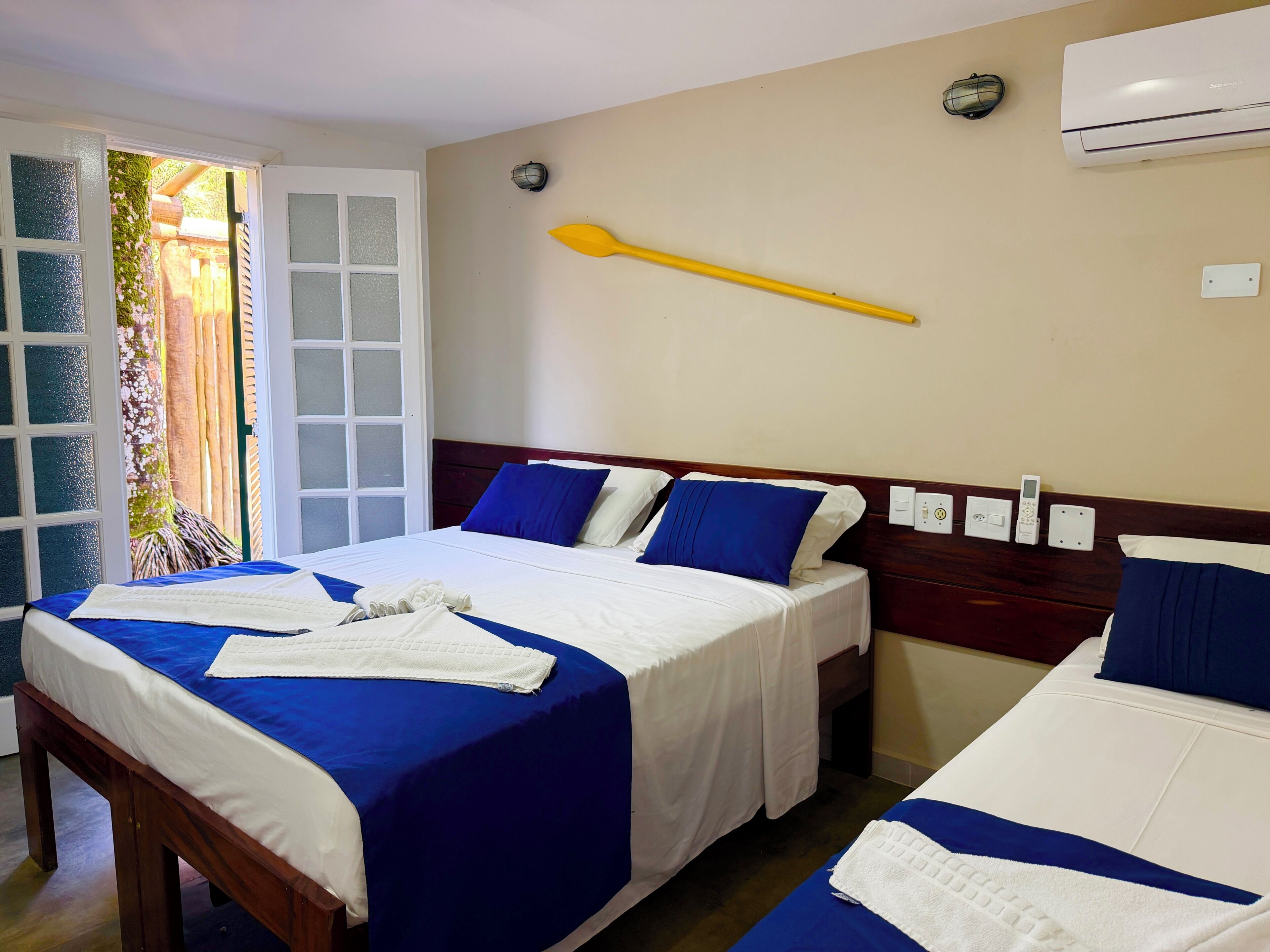 A hotel room with two beds, a wooden headboard, a wall-mounted air conditioner, and a window with a view of greenery.