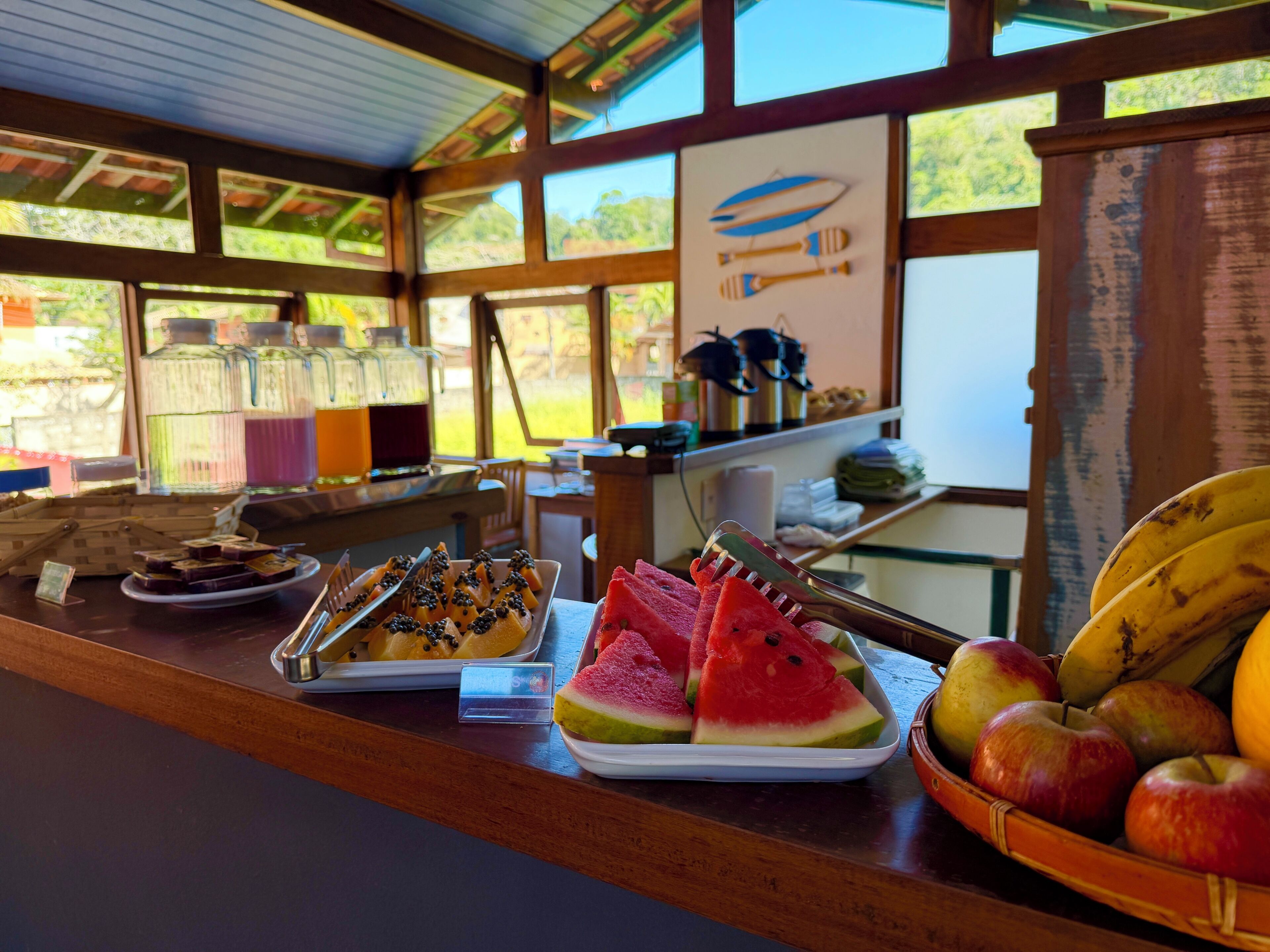 A breakfast buffet with various fruits and dishes, a wooden counter, and a view of the outdoors through large windows.