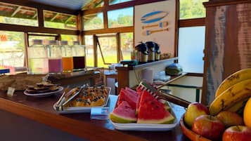 A breakfast buffet with various fruits and dishes, a wooden counter, and a view of the outdoors through large windows.