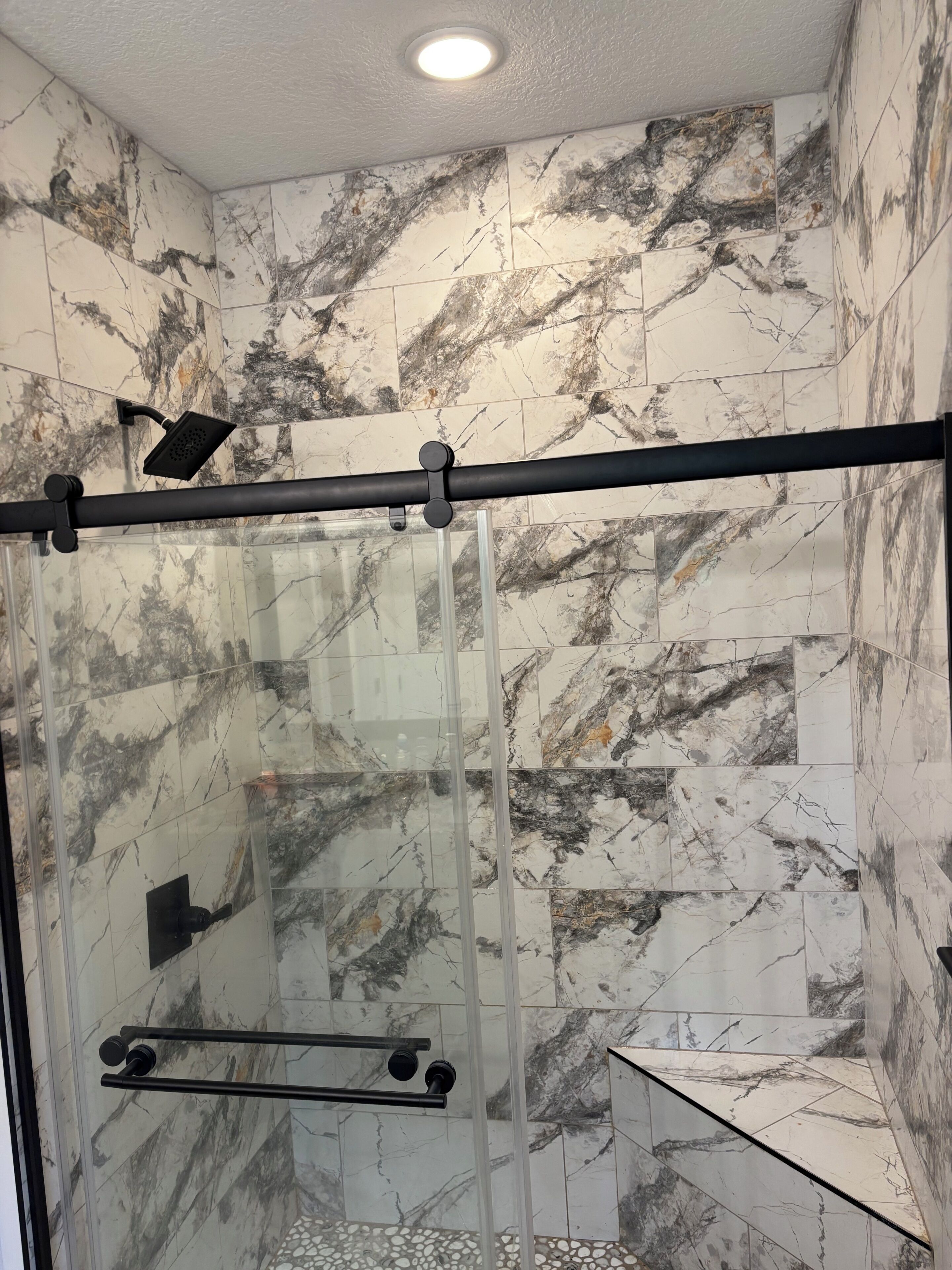 Een douche met glazen deur, marmeren tegels en een zwarte horizontale stang.