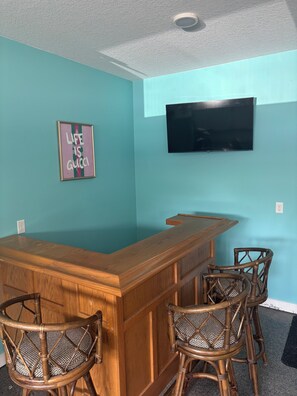 Una zona de bar con mostrador de madera y sillas de mimbre, un televisor de pantalla plana montado en la pared y un cartel enmarcado.