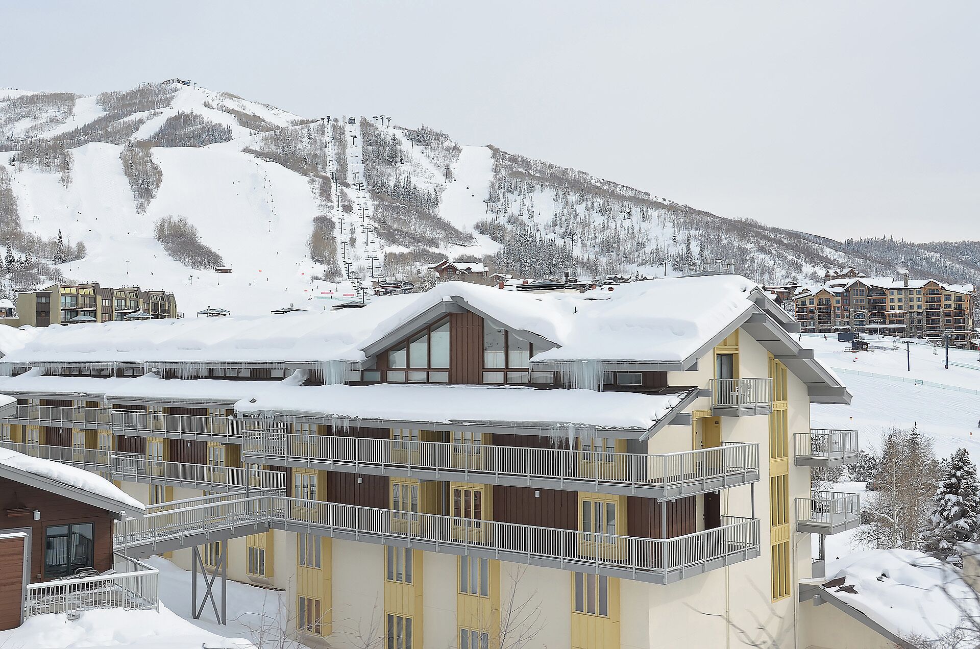 Un hôtel à plusieurs étages doté de balcons, avec un télésiège visible au loin et des montagnes enneigées.