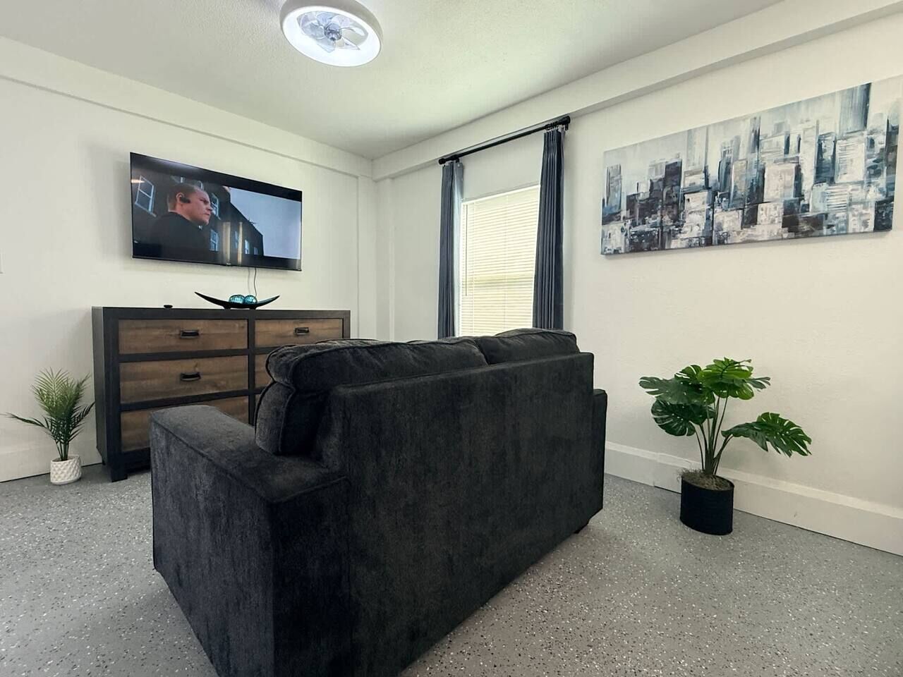 A modern living room with a dark sofa, a flat-screen TV mounted on the wall, a wooden cabinet, and a cityscape painting.