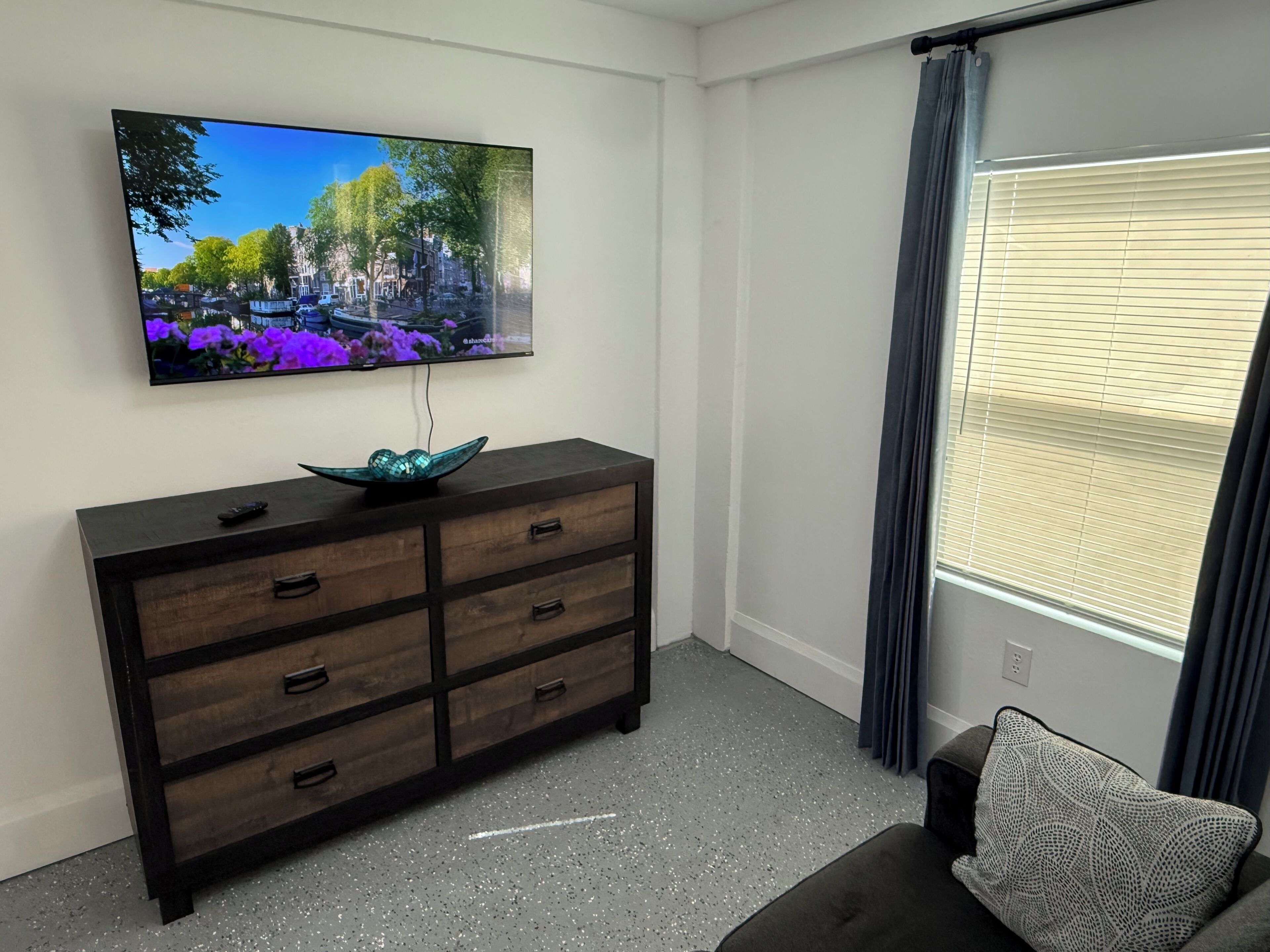 Una habitación con una televisión de pantalla plana montada en la pared, un aparador de madera oscura con cajones, una alfombra gris con motas y una ventana con persianas.
