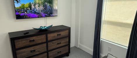 A room with a flat-screen TV mounted on the wall, a dark wooden dresser with drawers, a speckled grey carpet, and a window with blinds.