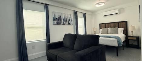 A hotel room with a bed, a sofa, a nightstand, and a wall-mounted air conditioning unit.