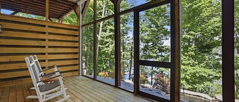 A wooden deck with a rocking chair and a bench, large windows offering a view of trees and a body of water.