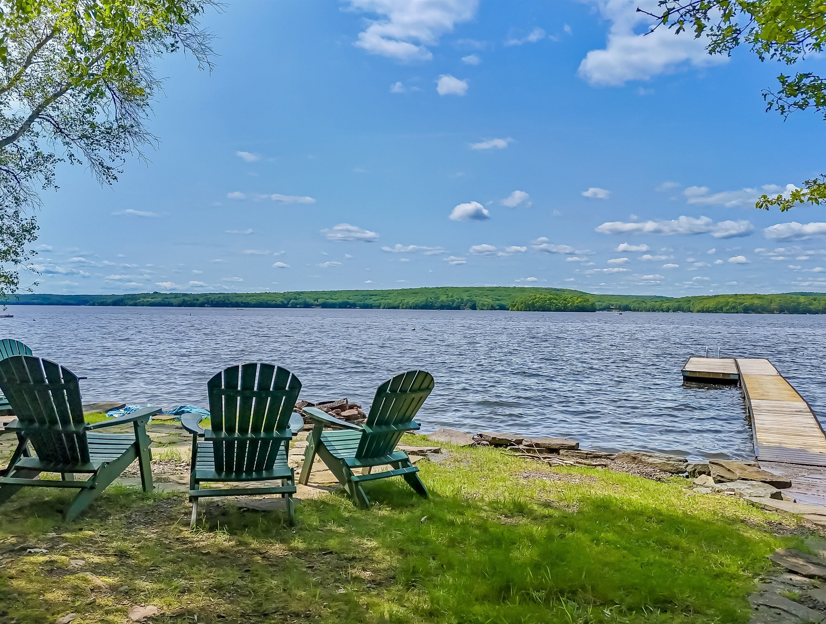 Blick auf ein Seeufer mit drei Adirondack-Stühlen, die zum Wasser zeigen, einer Holzsteganlage, die sich ins Wasser erstreckt, und einer Wiese im Vordergrund.