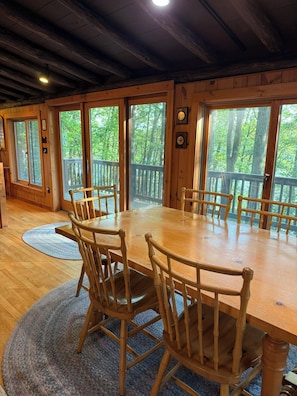 A wooden dining table with six chairs, large windows, and a view of trees.