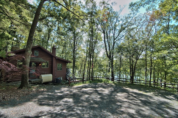 A rustic cabin with a gravel driveway and a wooden fence, surrounded by dense forest.